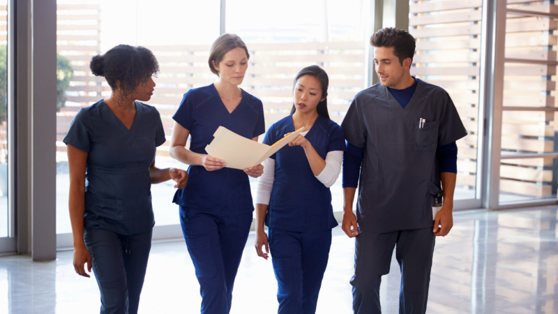 Healthcare,Colleagues,Discussing,Notes,In,Hospital,Corridor