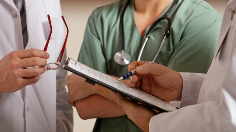 Midsection,Of,Doctor,Holding,Clipboard,While,Standing,With,Colleagues