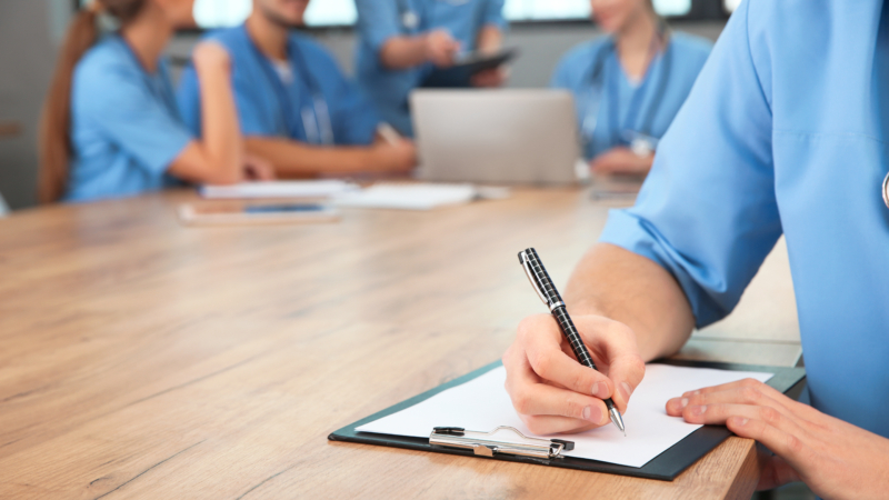 Medical,Student,With,Clipboard,And,His,Groupmates,Studying,In,University