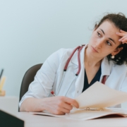 Tired,Female,Doctor,Sitting,At,Desk,Thoughtfully,Looks,Away,Holds