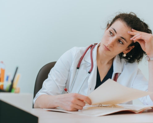 Tired,Female,Doctor,Sitting,At,Desk,Thoughtfully,Looks,Away,Holds