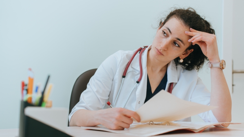 Tired,Female,Doctor,Sitting,At,Desk,Thoughtfully,Looks,Away,Holds