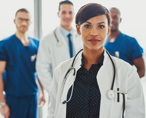 Confident,Female,Doctor,In,Front,Of,Team,,Looking,At,Camera