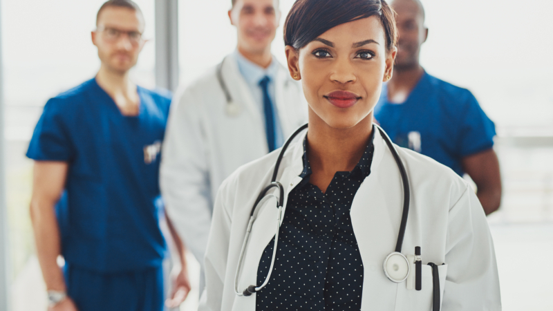 Confident,Female,Doctor,In,Front,Of,Team,,Looking,At,Camera